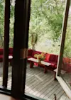 View through an open window of a wooden deck with red cushioned benches, wooden tables, and a bouquet of flowers, surrounded by lush green trees.