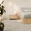 A bed with grey bedding sits next to a light wood nightstand with a lamp and books. A blurred plant is in the foreground.