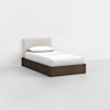 A single bed with a white upholstered headboard, white bedding, and a dark brown wooden base, placed in a minimalist, empty room with white walls and a light gray floor.