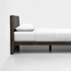 A side view of a minimalist bed with a dark wooden frame, a padded gray headboard, white bedding, and a pillow, set against a plain white wall and floor.