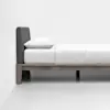 A minimalist bed with a wooden frame, dark upholstered headboard, white mattress, white pillow, and white bedding is placed against a white wall in a bright, empty room.