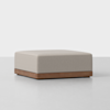 A square, light gray upholstered ottoman with a wooden base, placed on a smooth, light-colored floor against a plain white background.