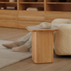 A person wearing light socks and pants sits on a cream-colored, cozy chair next to a wooden side table with a cup and saucer on top, in a room with wooden floors and shelves.