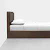 A modern wooden bed with a padded brown headboard, white bedding, and a single white pillow, shown in profile against a plain white wall and gray floor.