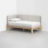 A minimalist wooden daybed with a beige upholstered backrest and single white mattress and pillow, positioned in the corner of a room with white walls and a light gray floor.