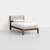 A single bed with a wooden frame, angled legs, a light gray upholstered headboard, and white bedding, positioned against a plain white wall on a light gray floor.