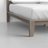 Close-up view of the corner of a wooden bed frame with light brown legs and a white mattress set against a light gray floor and white wall.