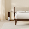 A modern bedroom with a wooden bed frame and beige upholstered headboard, white bedding, a wooden nightstand holding a glass carafe, two glasses, and several books, all set against a cream-colored wall and curtain.