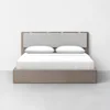 A modern, minimalist bed with a light wooden frame, a padded gray upholstered headboard, and white bedding with two pillows, set against a plain white wall and floor.