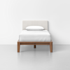 A single bed with a white upholstered headboard, white bedding, and a pillow, set on a wooden frame against a plain white wall and light gray floor.