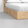 Close-up view of a modern wooden bed frame with white bedding and pillows, positioned on a smooth light gray floor next to a white wall.