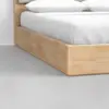 Close-up view of a modern wooden bed frame with white bedding and pillows, positioned on a smooth light gray floor next to a white wall.