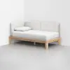 A minimalist daybed with a light wood frame, white mattress, single white pillow, and beige upholstered backrest, placed in the corner of a white room with a smooth gray floor.