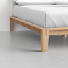 Close-up view of a wooden bed frame with light wood finish and clean lines, supporting a mattress with white bedding, set on a smooth gray floor against a white wall.