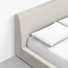 Close-up of a beige upholstered bed frame with a tall, padded headboard, neatly made with white pillows and sheets, placed on a light gray floor against a white wall.