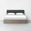 A neatly made bed with a black upholstered headboard, two white pillows, and white bedding, set in a light wood frame against a plain white wall and floor.