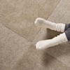 A person wearing light-colored socks and gray pants sits on a beige carpet made up of square tiles. Only their feet and lower legs are visible in the image.
