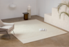 A modern, minimalist living space with a light beige rug, a gray armchair, a small brown side table, a white vase on a ribbed pedestal, an open book on the rug, and a plant by white steps against a neutral wall.