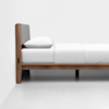 A modern wooden bed frame with a padded gray headboard, neatly made with white bedding and a single pillow, set against a plain white wall and floor.