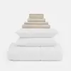 A neatly stacked bedding set includes white pillows and a comforter at the bottom, with folded beige sheets and pillowcases arranged in ascending size on top.