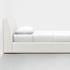 A minimalist side view of a modern, upholstered bed with a low headboard, white bedding, and a single pillow, set against a plain white wall on a gray floor.