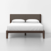 A modern bed with a dark wood frame and upholstered headboard, dressed in white bedding and two pillows, is centered against a plain white wall on a light gray floor.