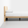 A minimalist wooden bed frame with a white mattress, white pillow, and white bedding, positioned against a white wall in a simple, bright room with a gray floor.