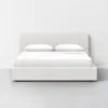 A modern, minimalist bed with a light beige upholstered frame and headboard, neatly made with white bedding and two stacked white pillows, set against a plain white wall on a gray floor.