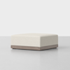 A modern, square ottoman with a cream-colored upholstered cushion and a wooden base, placed on a light gray floor against a plain white background.