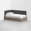 A modern daybed with a wooden base and dark gray upholstered back and side panels, placed in the corner of a minimalist white room, with a white pillow and neatly made white bedding.