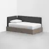A modern daybed with a wooden base and dark gray upholstered back and side panels, placed in the corner of a minimalist white room, with a white pillow and neatly made white bedding.