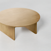 A minimalist, round wooden coffee table with a light finish, featuring a simple design and broad legs, set against a plain, light-colored background.