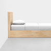 A minimalist wooden bed with a tall headboard, neatly made with white sheets and a pillow, positioned against a white wall on a light gray floor.