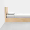 A minimalist wooden bed with a tall headboard, neatly made with white sheets and a pillow, positioned against a white wall on a light gray floor.