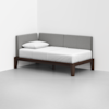 A modern daybed with a dark wooden frame, white mattress, single white pillow, and light gray upholstered backrest, positioned in the corner of a minimalist, white room.