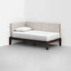 A modern daybed with a dark wooden frame, white mattress, single pillow, and light beige upholstered backrest, placed in the corner of a minimalist room with white walls and a gray floor.