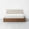 A minimalist bed with a light beige upholstered headboard, a white mattress labeled THUMA, and a wooden platform frame, set against a plain white wall and gray floor.