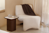 A cozy, white boucle lounge chair with a dark brown blanket draped over it sits next to a small wooden side table holding a teapot and cup, in a softly lit, neutral-toned room with sheer curtains.