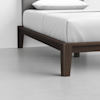 A close-up view of a modern bed with a dark wooden frame and legs, white bedding, and a gray upholstered headboard, set on a light gray floor against a plain white wall.