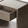 A minimalist, light wood cabinet stands on a plain white background. The cabinet has a single drawer, which is partially open, revealing the inside. The design is modern and simple.