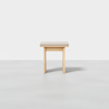 A small, minimalist wooden stool with a light-colored cushioned seat, photographed against a plain white background.