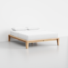 A minimalist wooden bed frame with a light finish, neatly made with white bedding and two white pillows, placed in a bright, empty room with a light floor and white walls.