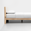 A minimalist wooden bed frame with white bedding and a single white pillow, positioned against a plain white wall on a light grey floor.