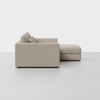 A modern, light beige sectional sofa with squared arms and backrests, viewed from the side, featuring an attached chaise lounge on the right. The background is plain white.