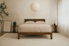 Minimalist bedroom with a wooden bed frame, beige bedding, matching nightstands, round paper lantern ceiling light, a potted plant, a vase, framed artwork on the floor, and soft natural light through sheer curtains.