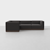 A modern, dark gray L-shaped sectional sofa with a low back and wooden base is set against a plain white background on a light gray floor.