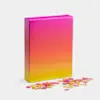 A rectangular puzzle box with a pink-to-yellow gradient design stands upright on a white surface. Several loose jigsaw puzzle pieces with matching gradient colors are scattered in front of the box.