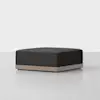 A modern, square black ottoman with a wooden base sits on a light gray floor against a plain white background.