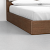 A close-up view of the corner of a wooden platform bed with white bedding and pillows, set on a smooth, light gray floor.