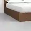 A close-up view of the corner of a wooden platform bed with white bedding and pillows, set on a smooth, light gray floor.